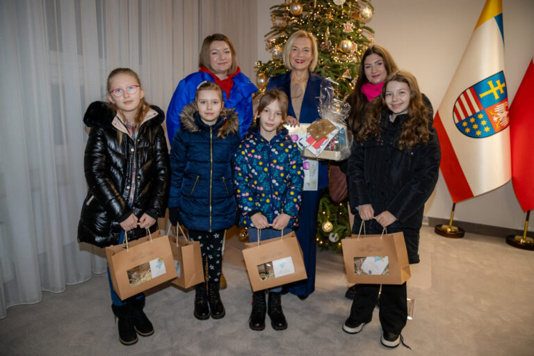 Dzieci z Klubu Wolna Strefa Jeziorańskiego podczas świątecznej wizyty, wraz z opiekunkami, w tle udekorowana choinka i flagi.