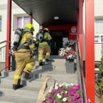Strażacy w sprzęcie ochronnym wchodzą do szkoły w Rykoszynie.
