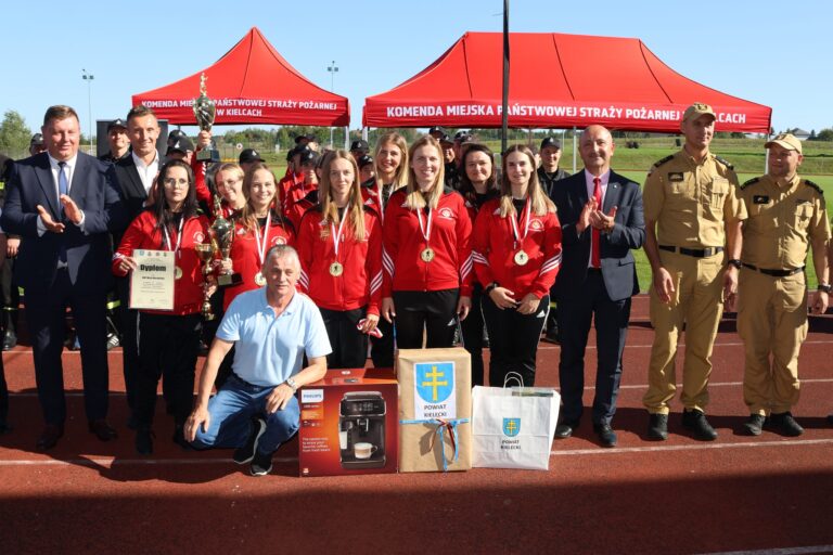 Zwycięska drużyna druhen i druhów na zawodach sportowo-pożarniczych, z pucharami i medalami, na tle namiotu straży pożarnej.