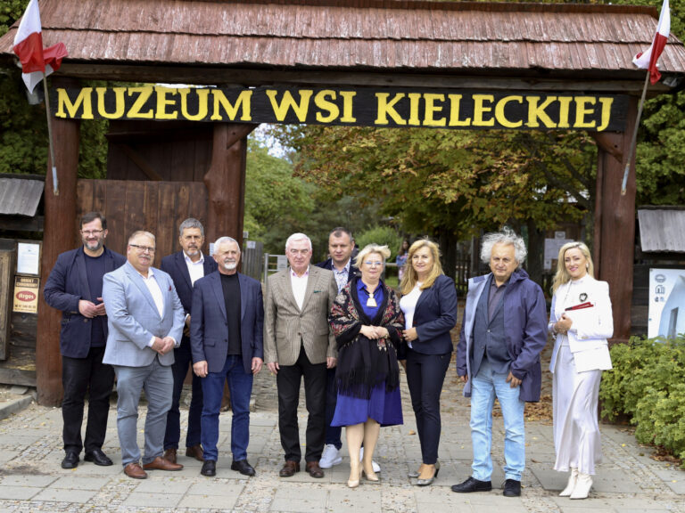 Grupa osób przed wejściem do Muzeum Wsi Kieleckiej, oznaczenia i flagi w tle.