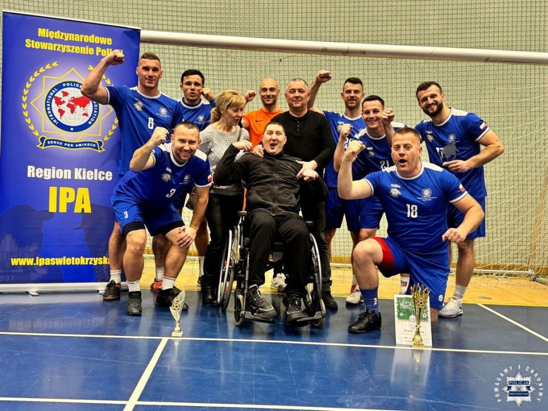 Grupa piłkarzy w niebieskich strojach oraz dwie osoby, w tym jedna na wózku inwalidzkim, pozują do zdjęcia na tle banera Międzynarodowego Stowarzyszenia Policji, Region Kielce IPA.