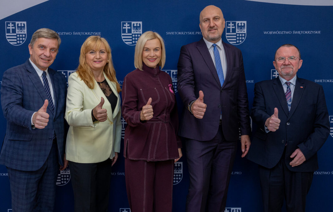 Grupa osób na tle banneru Województwa Świętokrzyskiego, pokazujących gest 'kciuki do góry'.