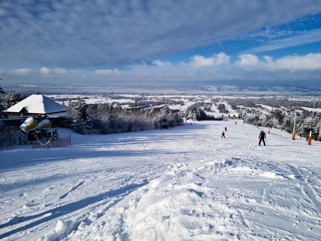 Zimowy krajobraz ze stokiem narciarskim oraz narciarzami na śniegu, wokół lasy i rozległe pola.