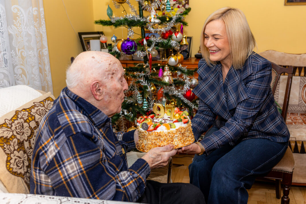 Starszy mężczyzna na kanapie z tortem urodzinowym, obok uśmiechnięta kobieta, w tle choinka
