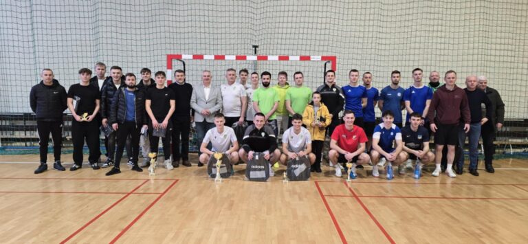 Grupa zawodników i organizatorów na Noworocznym Turnieju Piłki Halowej w Mniowie, stojących na hali sportowej przed bramką.