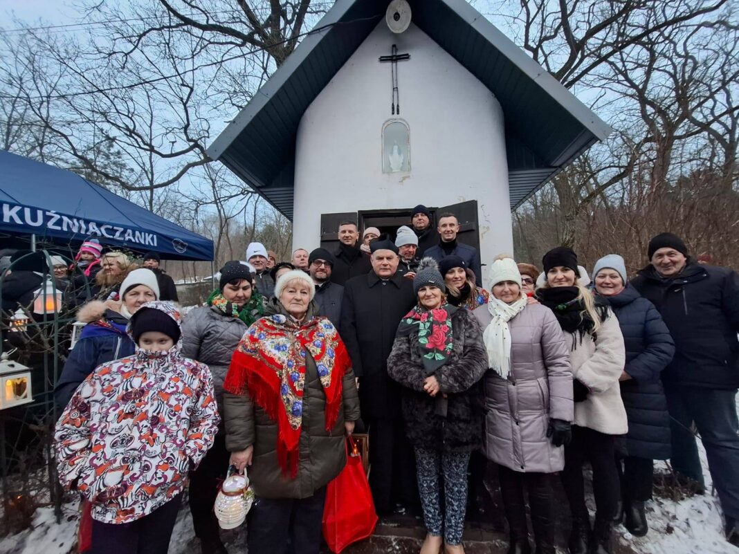 Grupa osób stojąca przed kapliczką podczas wydarzenia kolędowego w Kuźniakach.