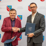 Dwie osoby ściskające dłonie na tle logo Gmina Nowiny, trzymające teczki z dokumentami.