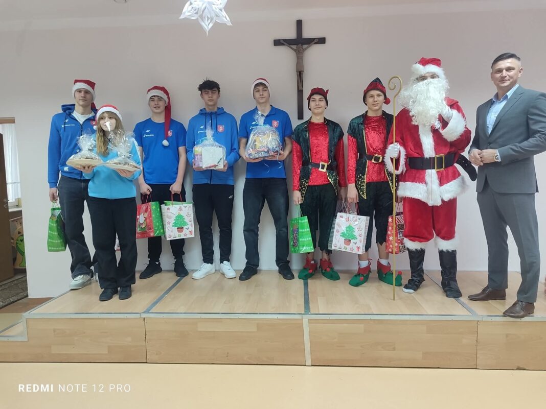 Grupa młodych ludzi w świątecznych strojach na scenie podczas wizyty w Domu Pomocy Społecznej.