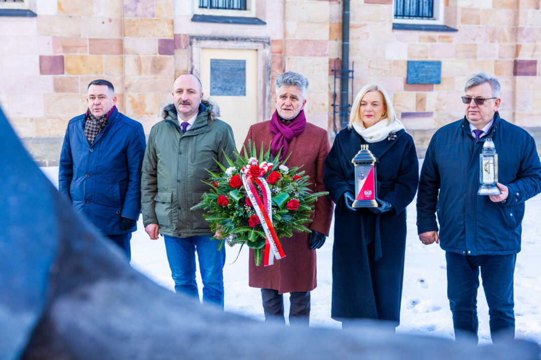 Grupa osób składająca hołd z kwiatami i zniczami przed budynkiem