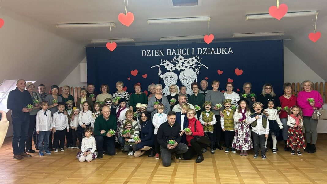 Grupa dzieci i dorosłych podczas obchodów Dnia Babci i Dziadka w Przedszkolu Samorządowym w Morawicy, na tle ozdobnej dekoracji