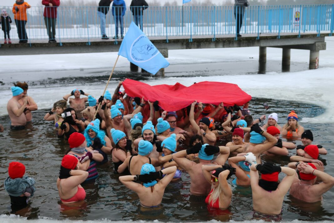 Grupa ludzi w czapkach kąpie się w zimowym jeziorze, trzymając czerwoną płachtę i niebieską flagę.