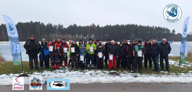Grupa uczestników w zawodach wędkarskich podlodowych nad zalewem w Morawicy, stojących na pokrytym śniegiem terenie z lasem w tle.