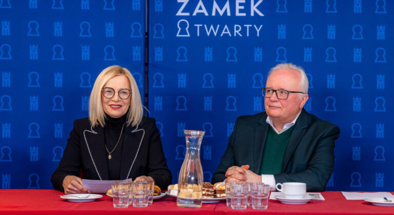 Dwie osoby siedzące przy stole na tle baneru 'Zamek Otwarty'