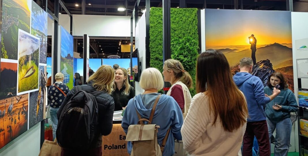 Stoisko targowe z wieloma osobami oglądającymi i rozmawiającymi. Na ścianach znajdują się duże zdjęcia prezentujące krajobrazy i atrakcje turystyczne.