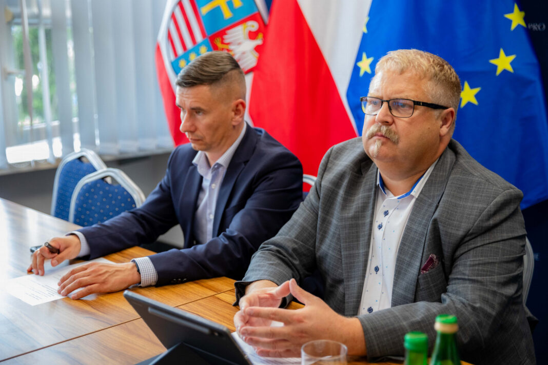 Dwóch mężczyzn podczas obrad komisji, polska flaga i unijna flaga w tle